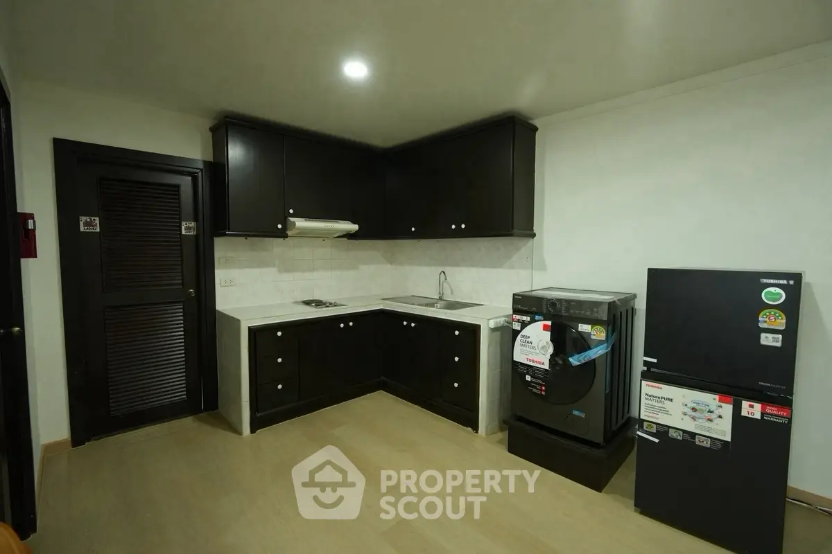 Modern kitchen with sleek black cabinets, washing machine, and compact fridge in a cozy apartment.