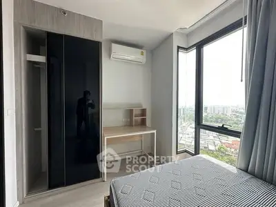 Modern bedroom with large window offering city view, featuring sleek wardrobe and compact desk.