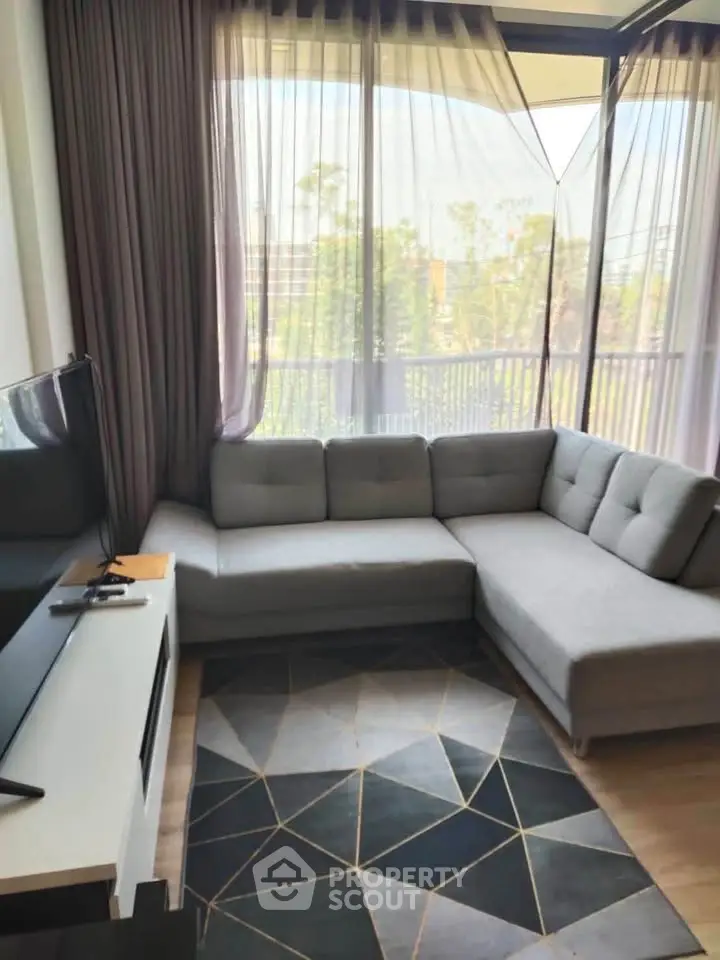 Modern living room with stylish gray sectional sofa and geometric rug, featuring large windows with sheer curtains.