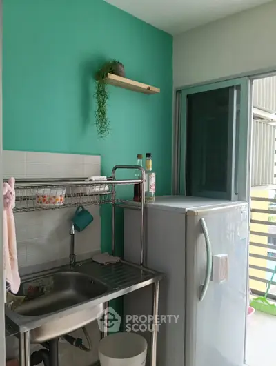Compact kitchen with stainless steel sink and fridge, featuring a vibrant teal accent wall and balcony access.
