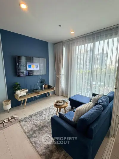 Modern living room with blue sofa, wall-mounted TV, and large windows offering city views.