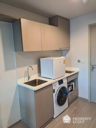 Compact modern kitchenette with built-in cabinets, washing machine, and fridge, perfect for urban living in a space-efficient apartment.