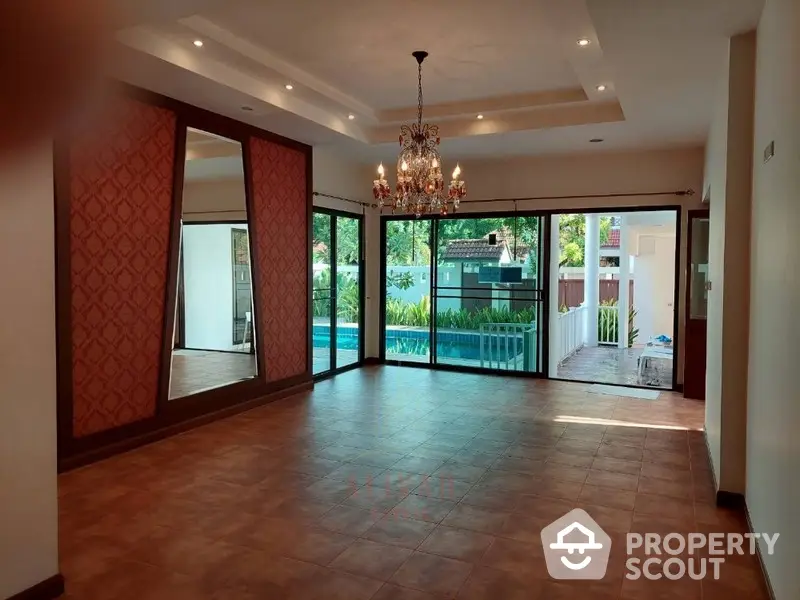 Spacious living room with elegant chandelier, large windows, and direct access to a serene pool area, perfect for entertaining and relaxation.