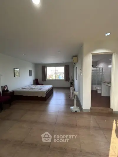 Spacious studio apartment with abundant natural light, featuring a large bed, ample floor space, and a glimpse into a tiled kitchen area.