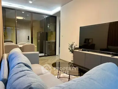 Modern living room with sleek furniture and large TV, open layout to kitchen area.