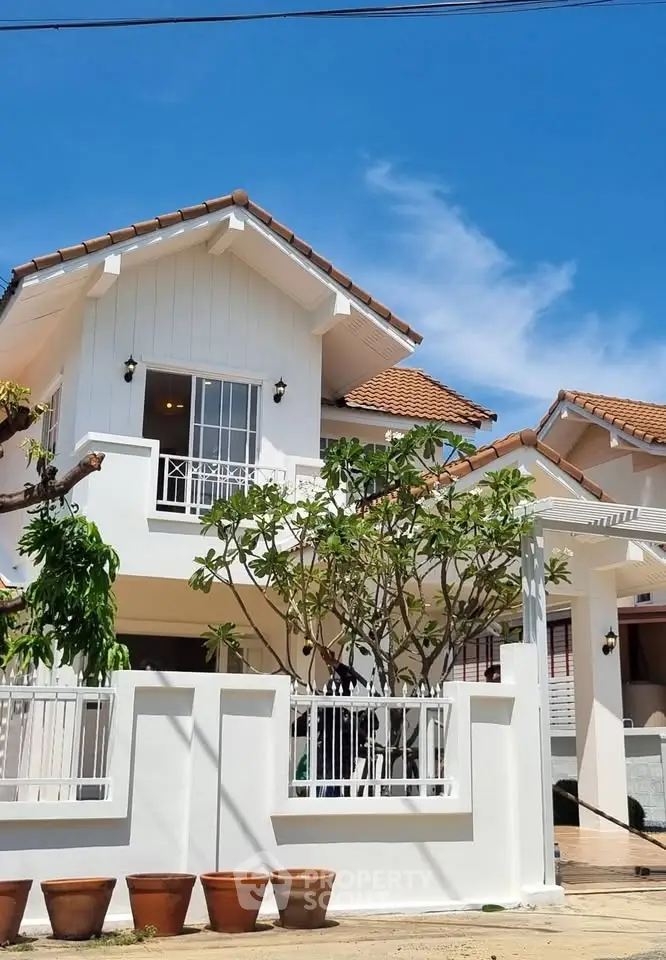 Charming two-story house with white facade and terracotta roof, surrounded by lush greenery and blue sky.