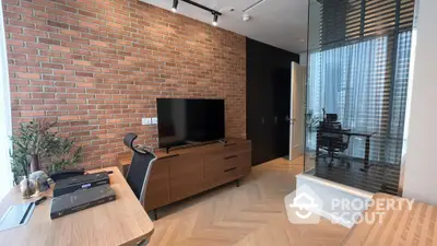 Chic urban living room with exposed brick wall, sleek entertainment unit, and herringbone wood flooring, leading to a modern glass-partitioned study.