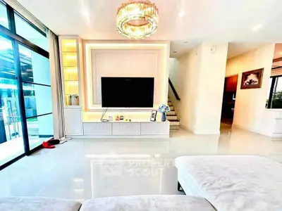 Luxurious modern living room with elegant chandelier and large TV, featuring sleek white decor and ample natural light.