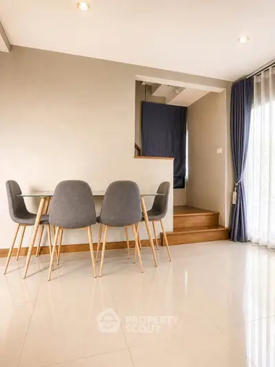Modern dining area with sleek glass table and stylish gray chairs, featuring natural light from large windows.