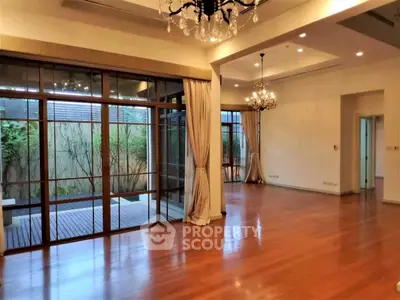 Spacious living room with hardwood floors and elegant chandeliers, featuring large windows overlooking a garden.