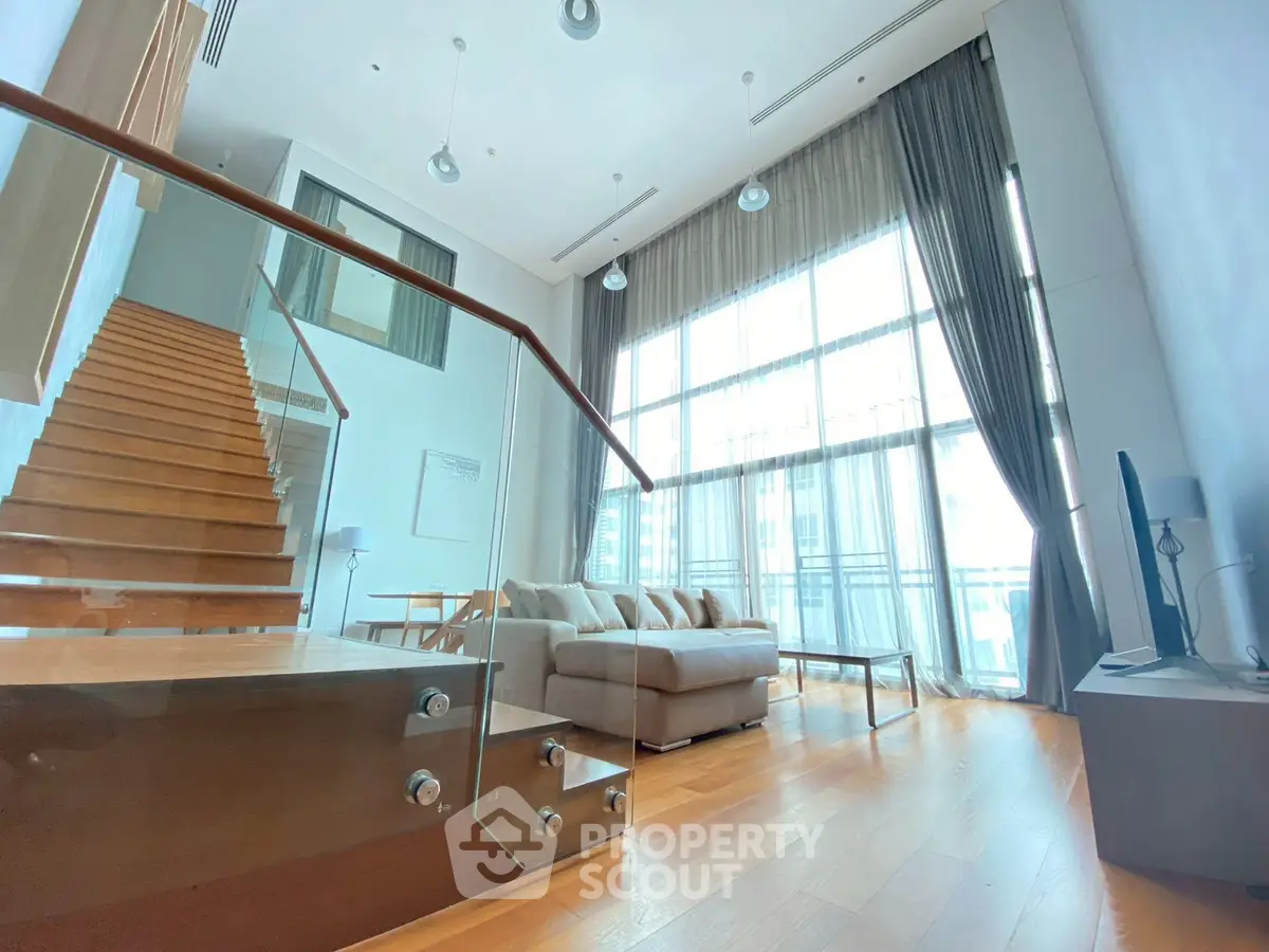 Spacious modern living room with high ceilings and large windows, featuring a stylish staircase and cozy seating area.