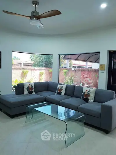 Spacious living room with modern sectional sofa and glass coffee table, featuring large windows and ceiling fan.