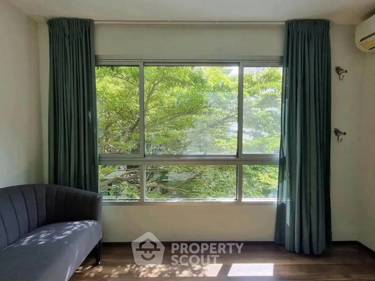 Bright living room with large window and lush green view, featuring elegant gray sofa and wooden flooring.