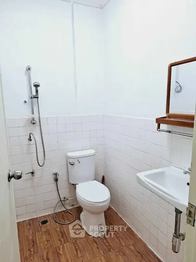 Compact bathroom with white tiles and wooden flooring, featuring a toilet, sink, and shower.