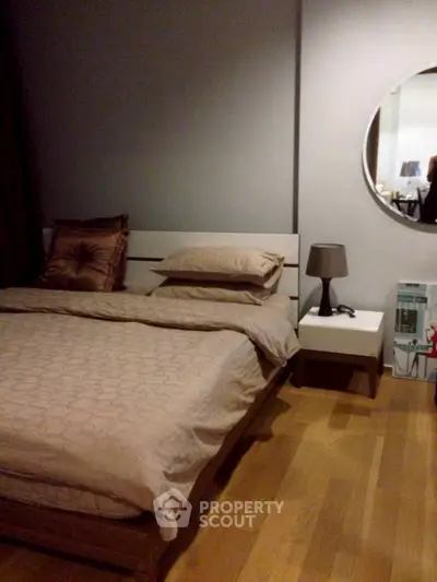 Cozy bedroom with modern decor and wooden flooring, featuring a stylish round mirror and elegant bedside lamp.