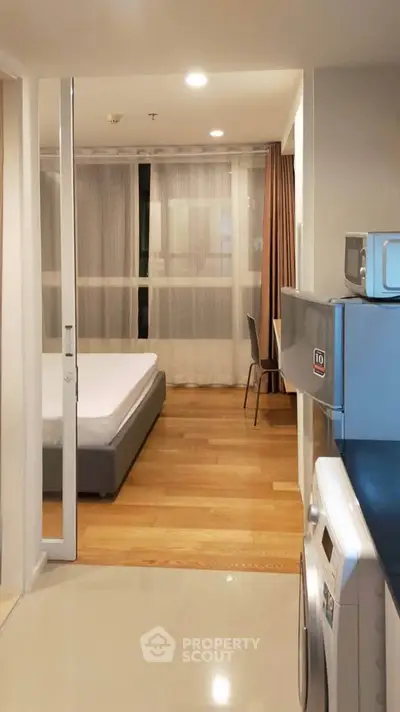 Modern apartment interior with bedroom and kitchen appliances, featuring wooden flooring and natural light.