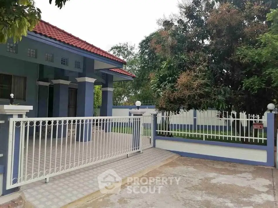 Charming blue house with red roof and spacious gated driveway, surrounded by lush greenery.
