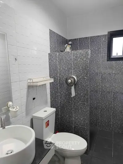 Modern bathroom with sleek gray tiles and white fixtures, featuring a compact shower area and window for natural light.