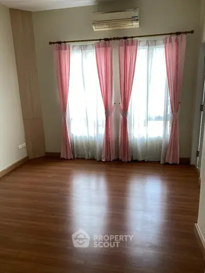 Spacious and well-lit empty room with polished wooden flooring, elegant pink curtains, and a modern air conditioning unit, ready for personalization.