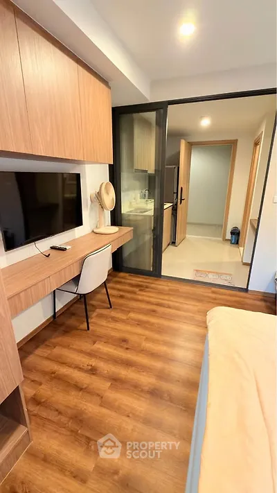 Modern bedroom with wooden flooring and study area, featuring a sleek kitchen view through glass doors.