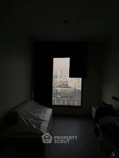 Dark room with a large window offering a cityscape view, featuring a covered sofa and minimal decor.