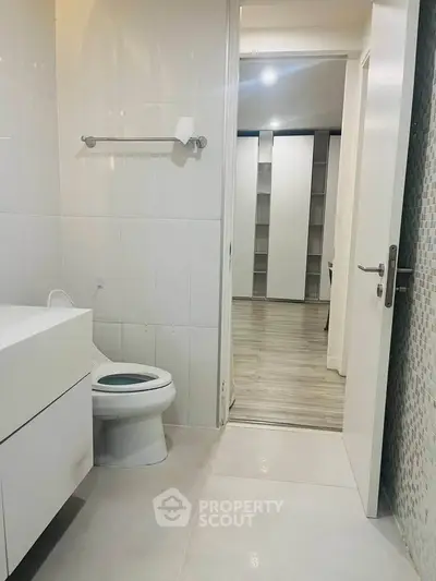 Modern bathroom with sleek white fixtures and view into adjacent room with wooden flooring.