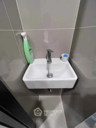 Compact bathroom sink with modern faucet and cleaning supplies in a tiled setting.