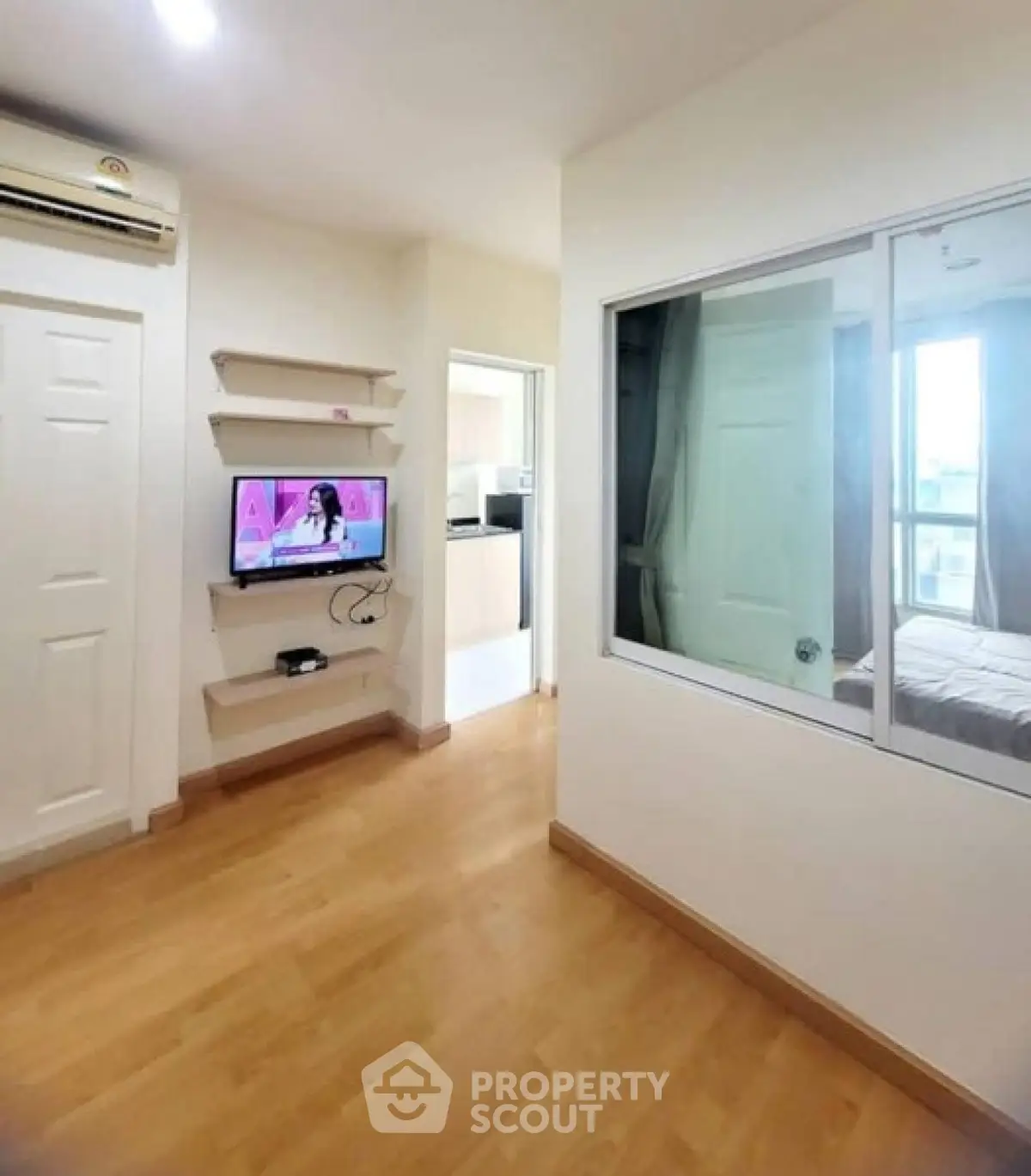 Modern apartment interior with wooden flooring, wall-mounted TV, and view into bedroom and kitchen.