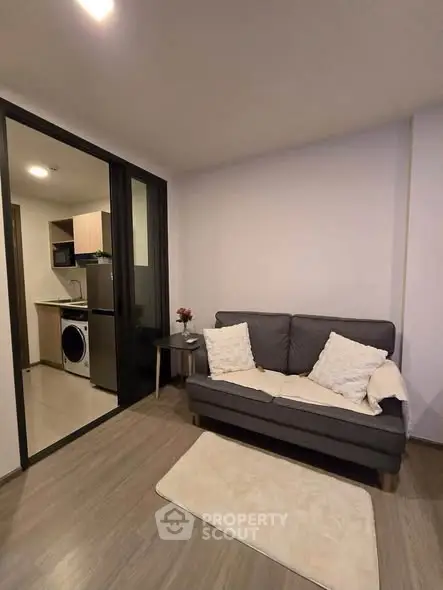 Modern living room with cozy sofa and open kitchen featuring washing machine and fridge.