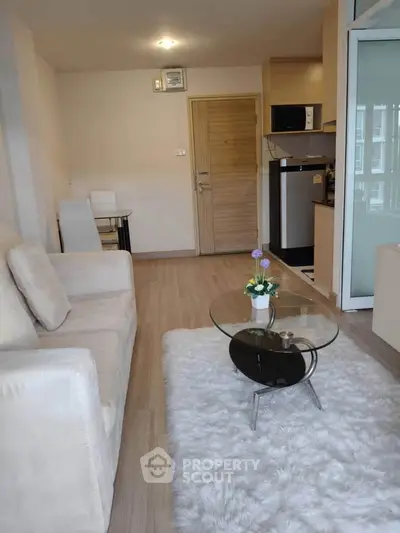 Modern living room with cozy white sofa and glass coffee table, adjacent to a compact kitchen.