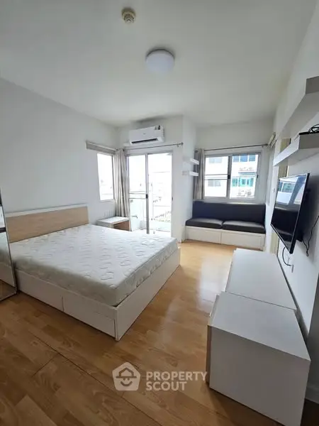 Modern bedroom with wooden flooring, large windows, and a wall-mounted TV in a bright apartment.