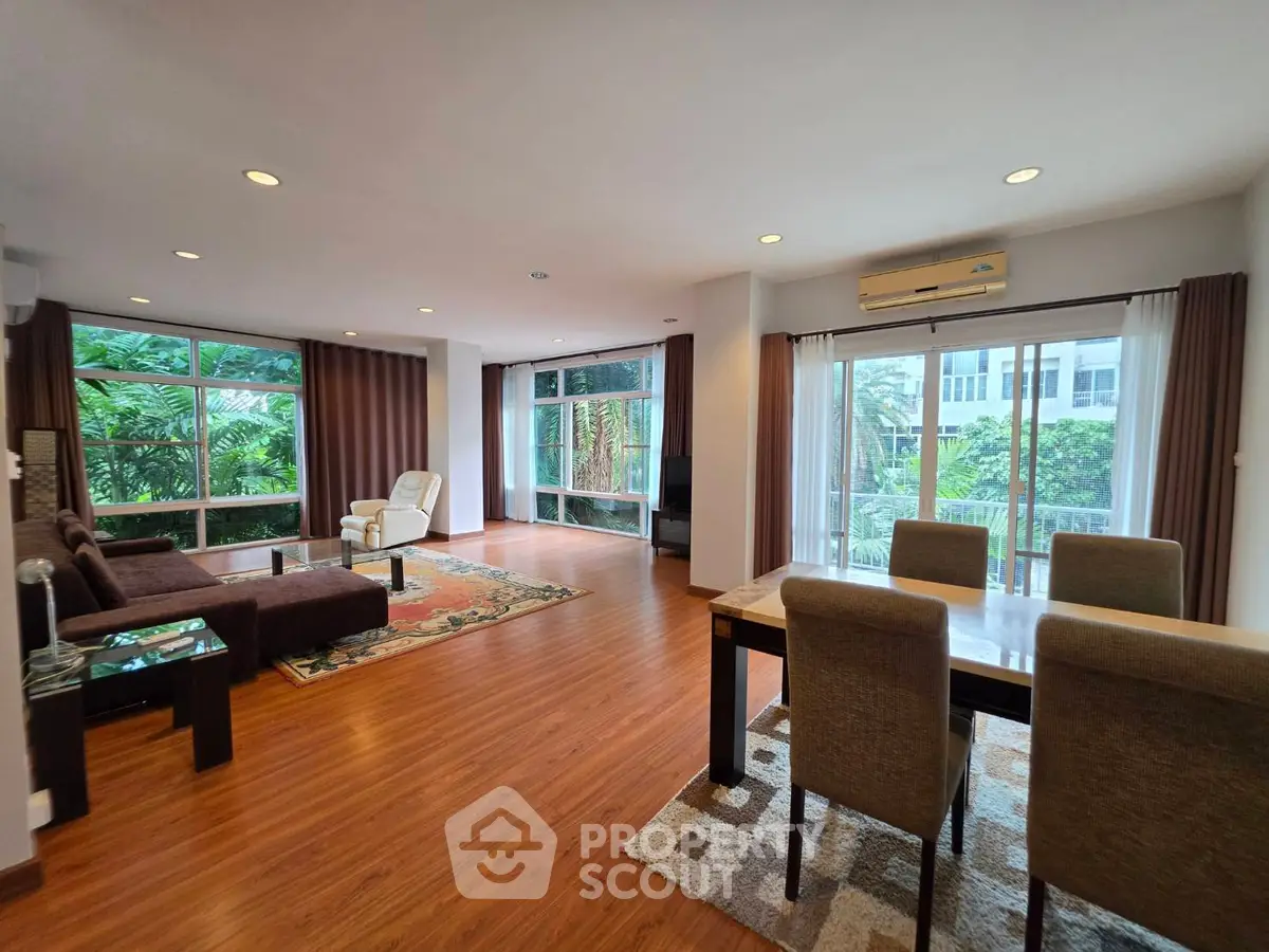 Spacious living room with large windows and garden view, featuring modern furniture and elegant wooden flooring.