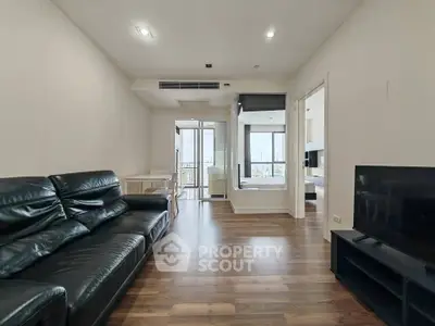 Spacious modern living room with sleek black leather sofa and wooden flooring, offering a glimpse into a bright bedroom.
