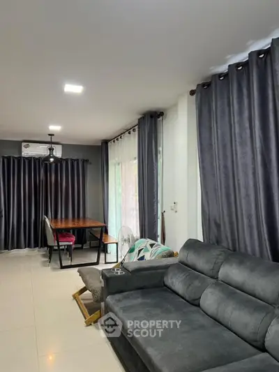Spacious living room with modern gray sofa and dining area, featuring elegant curtains and bright lighting.