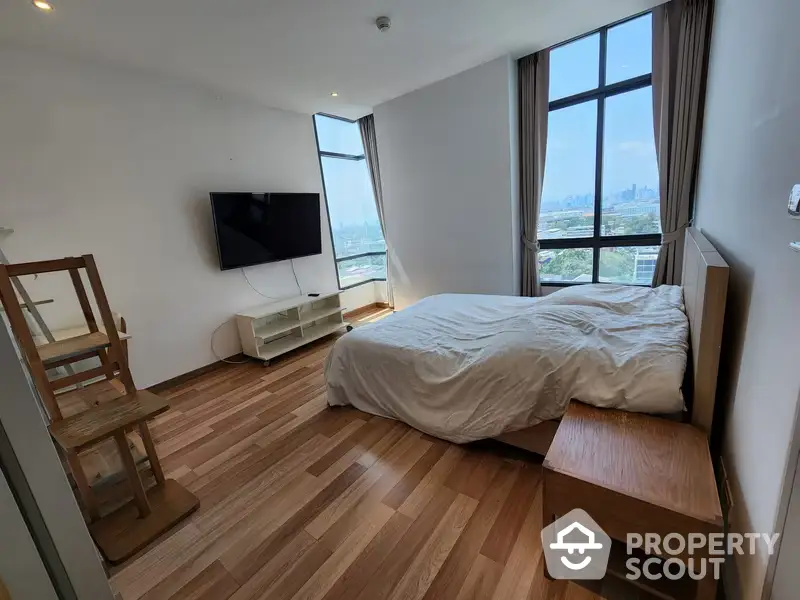 Bright and airy bedroom with large windows offering a panoramic city view, polished wooden flooring, and modern furnishings including a wall-mounted TV.