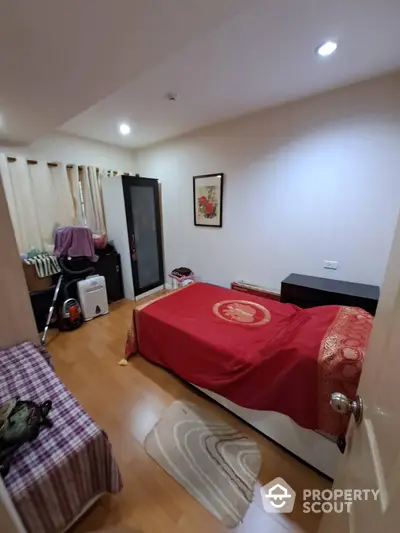 Spacious bedroom with polished wooden flooring, a large comfortable bed with a red cover, and ample lighting creating a warm ambiance.