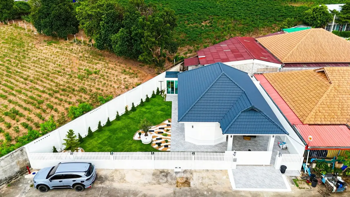 Aerial view of a modern house with a blue roof, spacious garden, and adjacent farmland.
