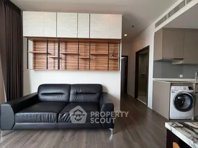 Modern living room with sleek black sofa and stylish wooden shelving, adjacent to a compact kitchen with washing machine.