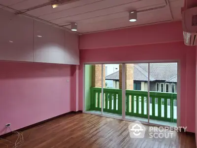 Spacious room with vibrant pink walls and hardwood flooring, leading to a charming balcony with a green railing, offering a suburban view.