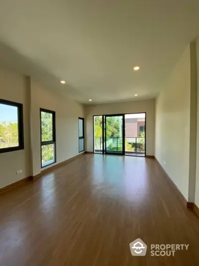 Spacious and sunlit living room with polished hardwood floors, large windows, and a glass door leading to a balcony, offering a serene tree-lined view.