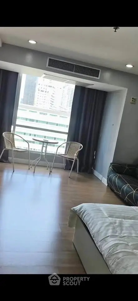 Modern studio apartment with large window and city view, featuring cozy seating area and sleek wooden flooring.