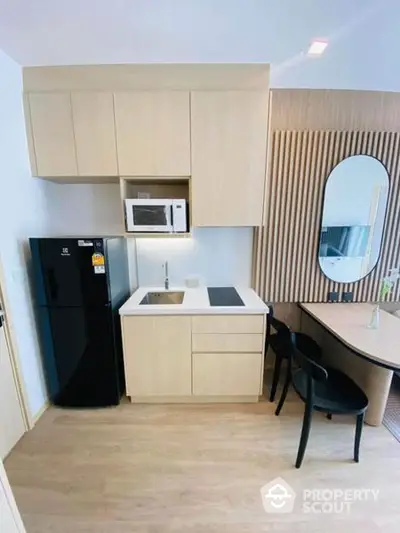 Compact modern kitchen with sleek beige cabinetry, black refrigerator, built-in microwave, and a cozy dining area with stylish chairs and oval mirror.