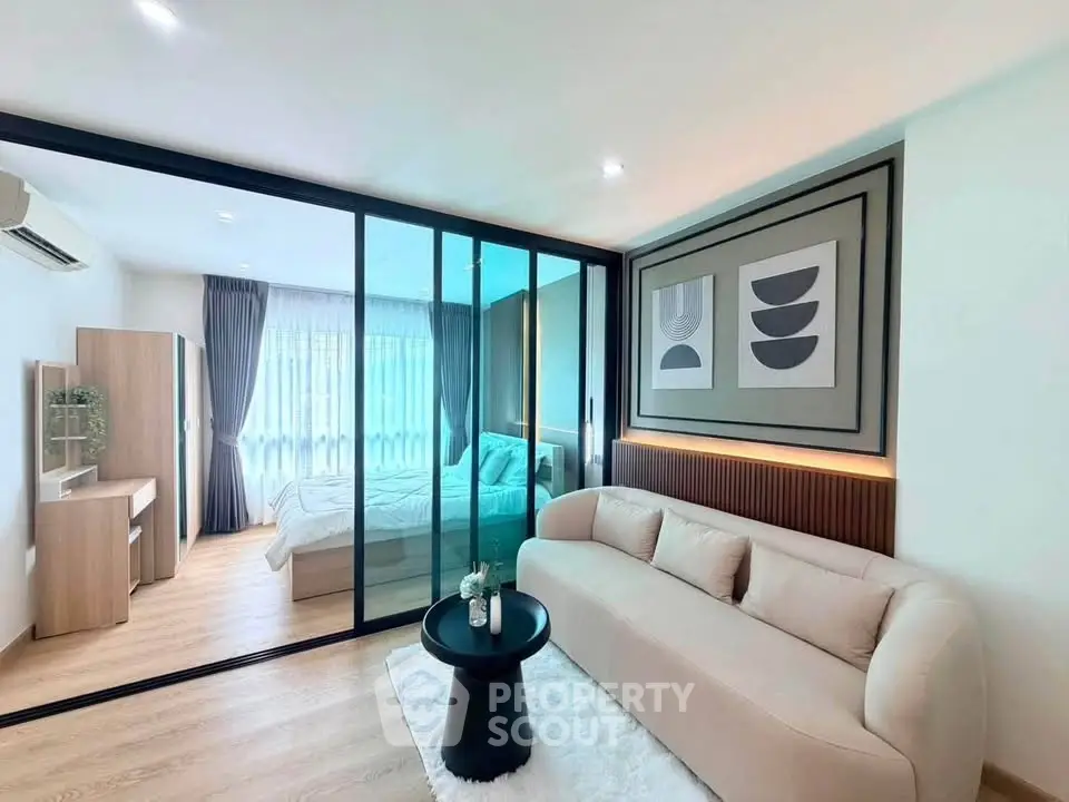 Modern living room with glass partition leading to a cozy bedroom, featuring stylish decor and natural light.