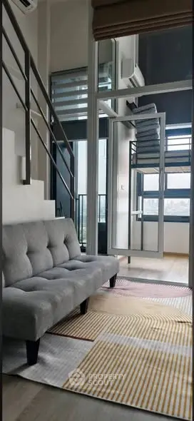 Modern loft apartment with stylish gray sofa and mezzanine bed, featuring large windows and a cozy ambiance.