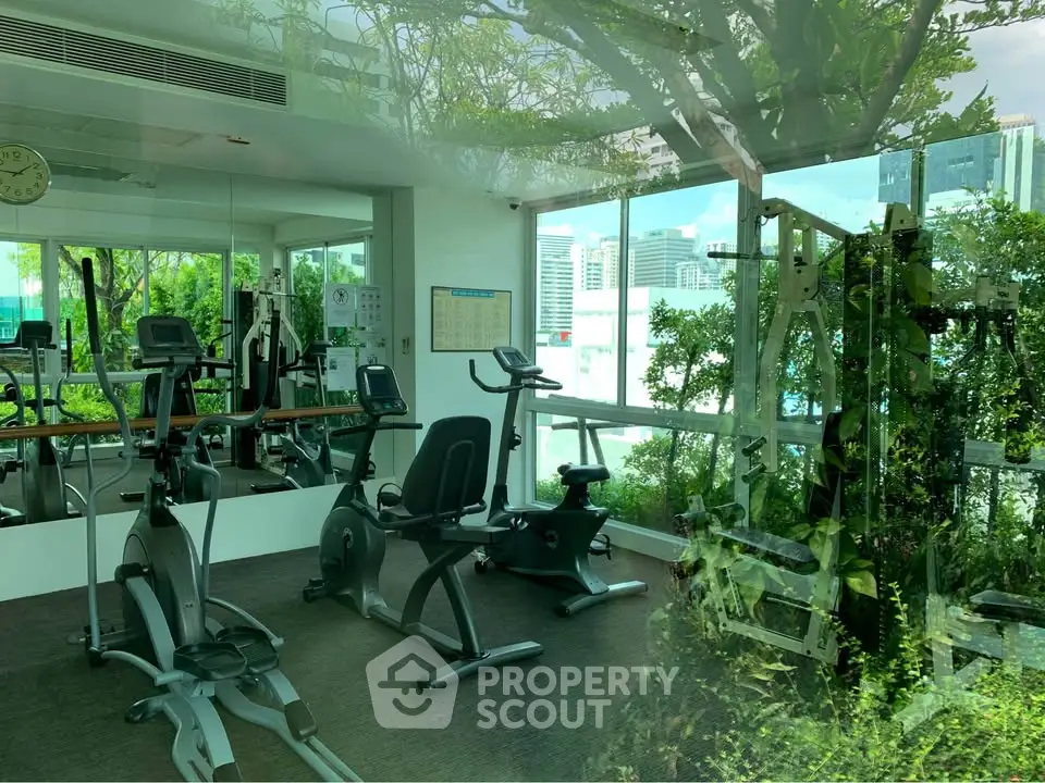 Modern gym with city view and lush greenery, featuring exercise bikes and fitness equipment.