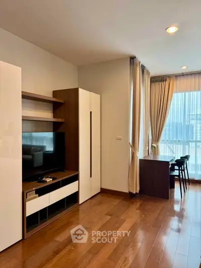 Modern living room with wooden flooring, TV unit, and large windows offering natural light.