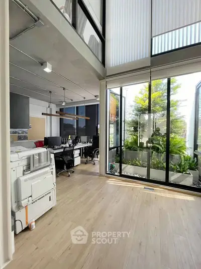 Modern office space with high ceilings and large windows overlooking a lush garden, featuring sleek furniture and natural light.