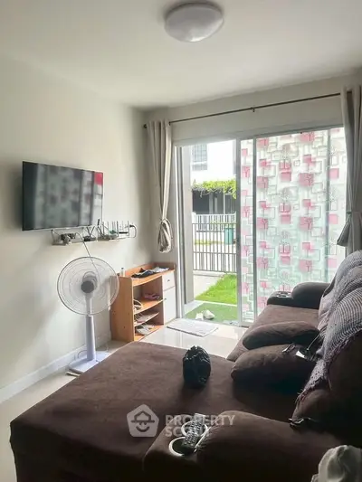 Cozy living room with brown sofa, wall-mounted TV, and sliding glass doors leading to a garden view.