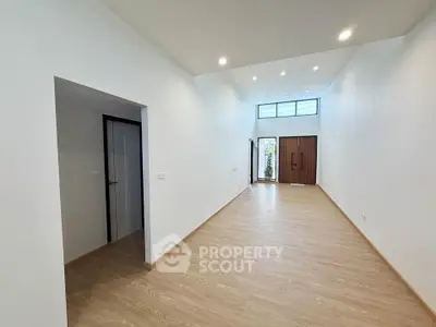Spacious modern living room with high ceiling and wooden flooring, featuring a large entrance door and natural light.