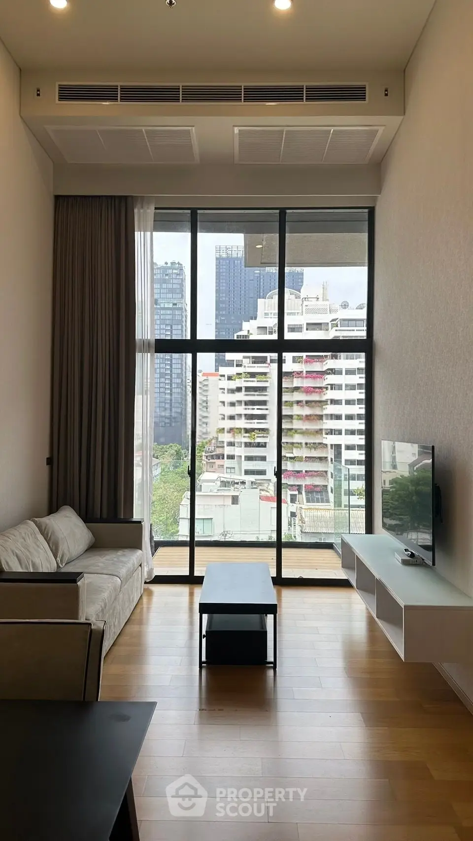 Modern living room with high ceiling and city view, featuring sleek furniture and large windows.